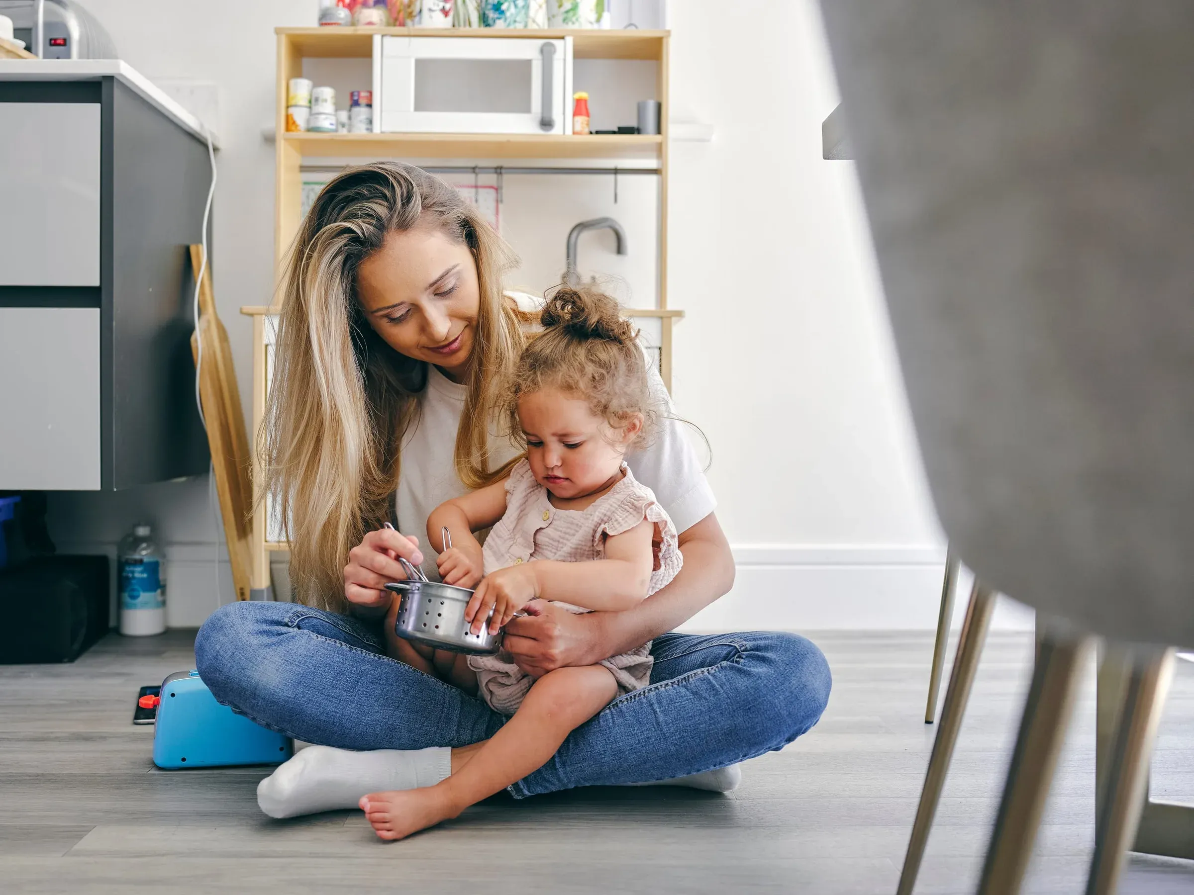 A mother and her toddler playing on the kitchen floor with a saucepan
