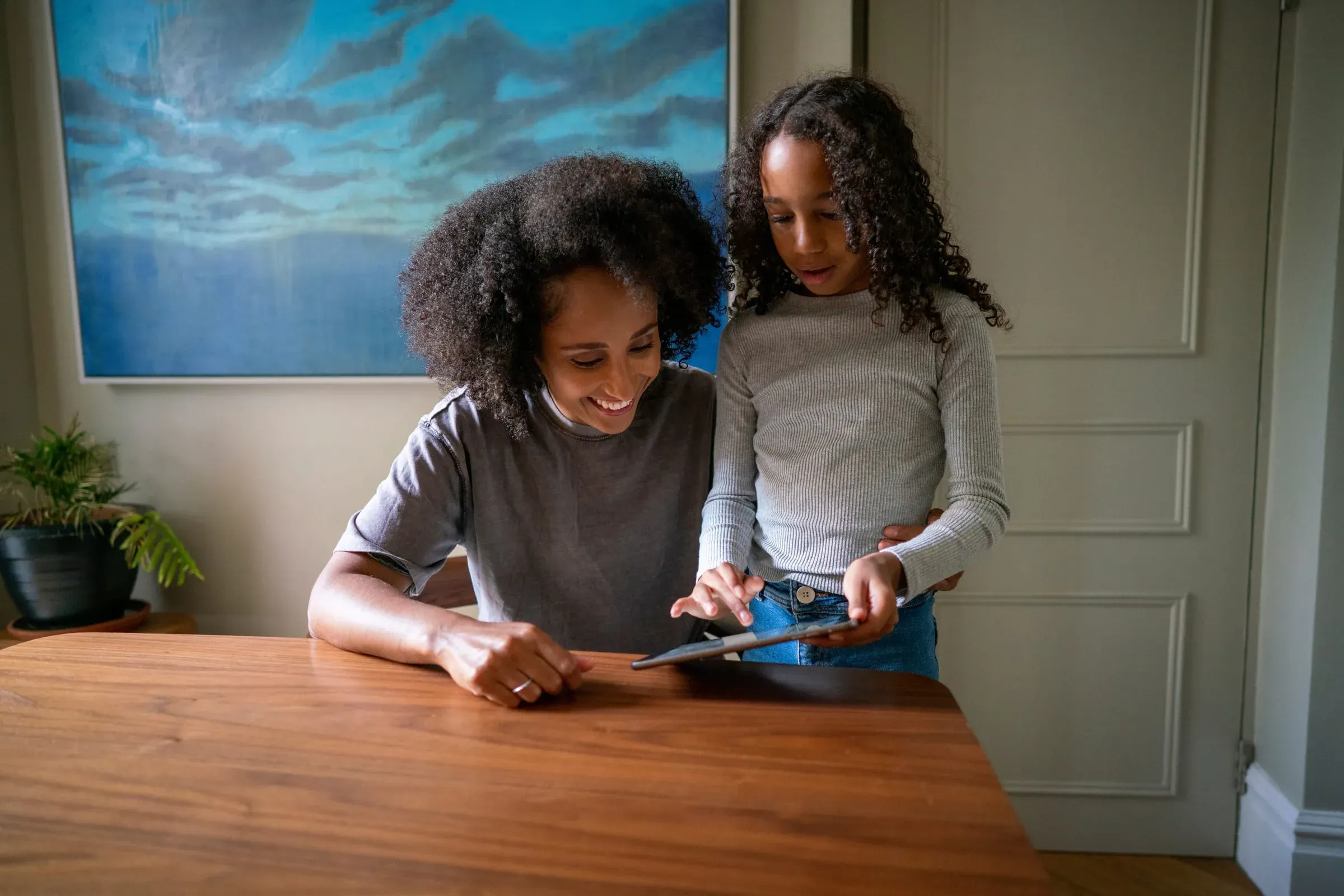 A mother and her young daughter at a kitchen table looking at a tablet together